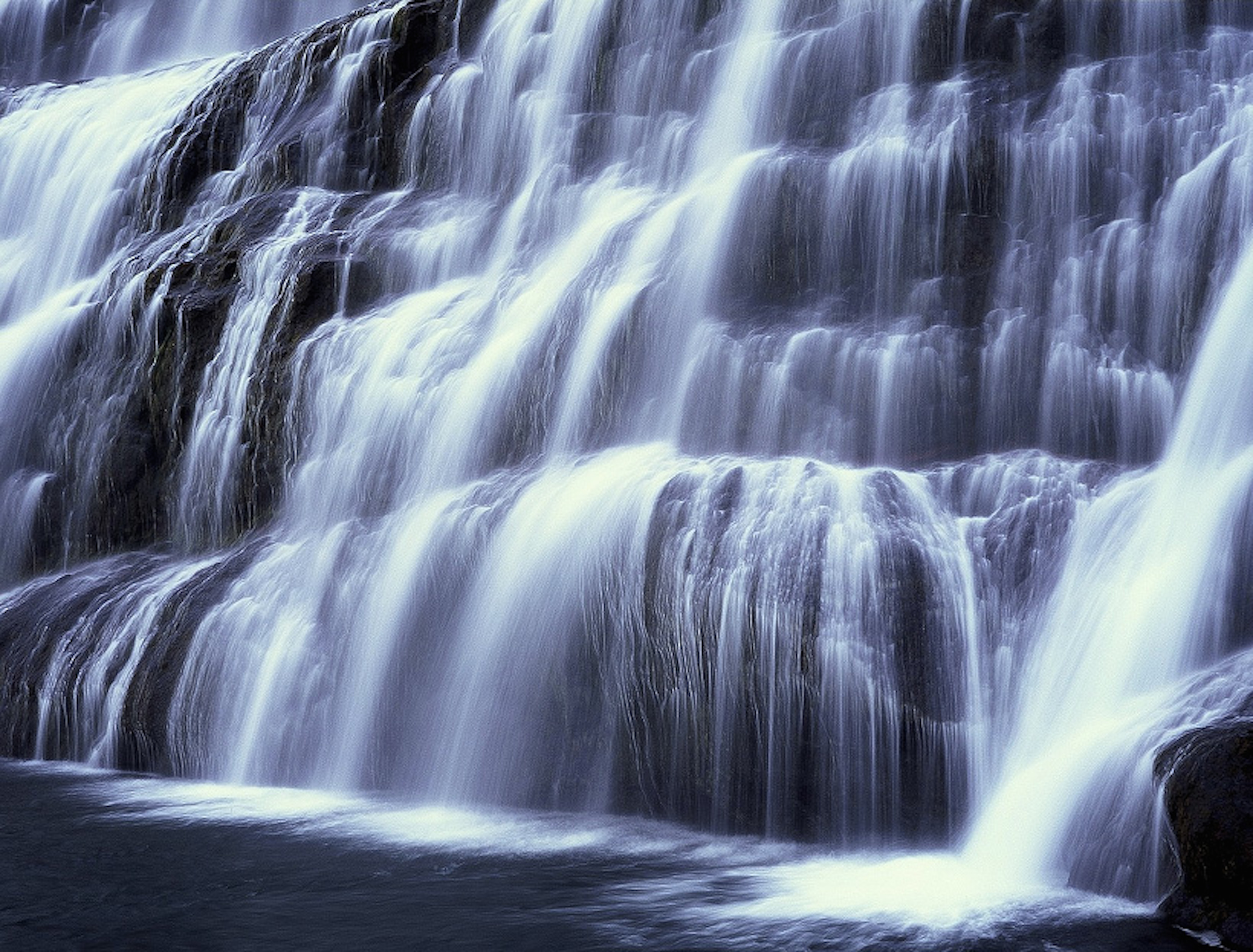 Waterfall wallpaper — long-exposure photograph of a multi-tiered cascade of white water over dark rocks in cool blue tones
